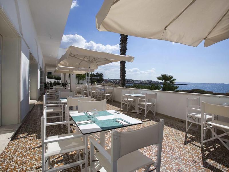 Terrasse mit weißen Tischen und Stühlen, Sonnenschirmen, Meerblick und blauem Himmel.