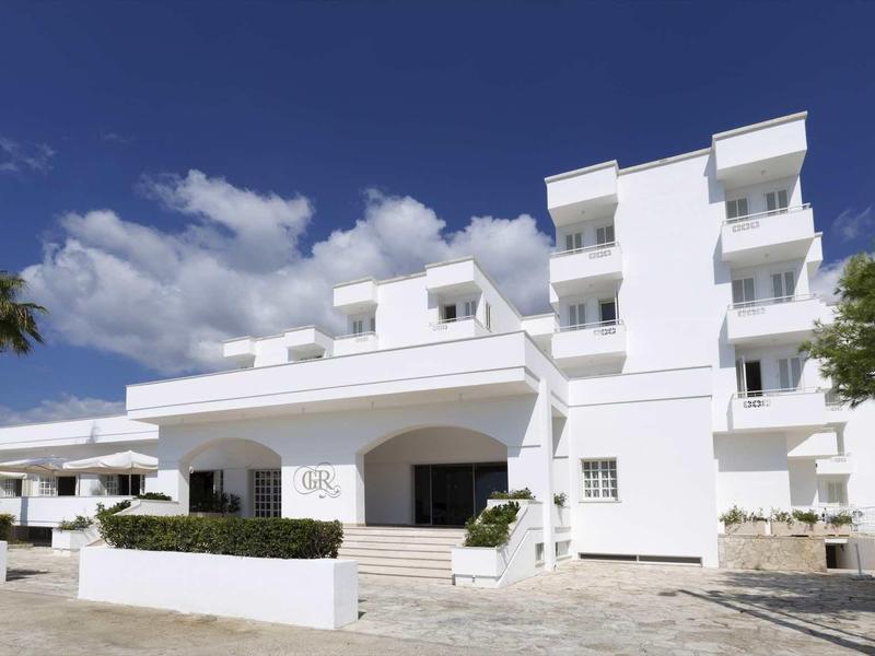 Modernes weißes Gebäude mit mehreren Stockwerken, Balkonen und Palmen vor blauem Himmel.
