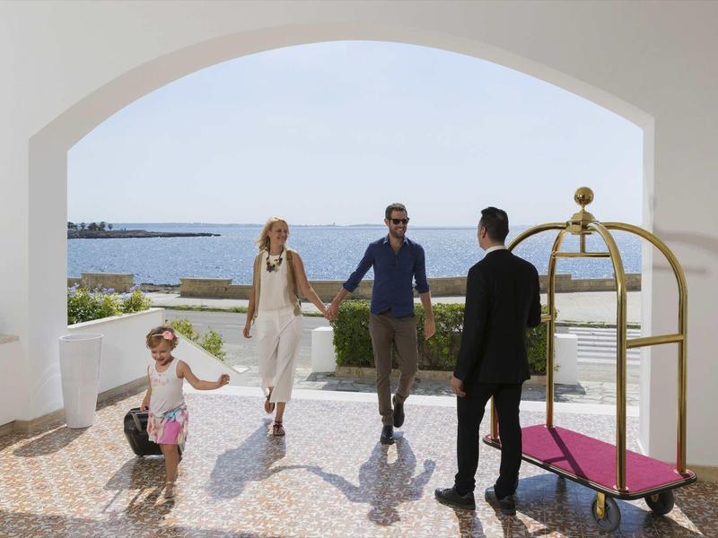 Vier Personen in heller, offener Hotellobby mit Blick aufs blaue Meer und gepflastertem Boden.