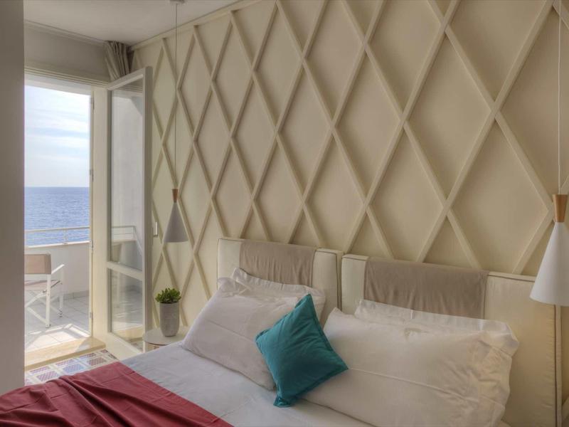 Helles Schlafzimmer mit weißem Bett, türkisfarbener Kissen und Balkon mit Meerblick.