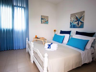 Hell eingerichtetes Schlafzimmer mit weißem Bett, blauer Deko und blauen Vorhängen am Fenster.