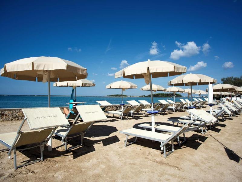 Strand mit weißen Sonnenschirmen und Liegestühlen, blauer Himmel und ruhiges Meer im Hintergrund.