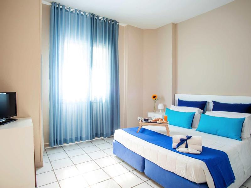 Helles Schlafzimmer mit blauer Bettwäsche, zwei blauen Kissen, Flachbildfernseher und Vorhängen am Fenster.