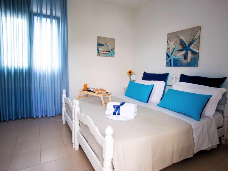 Hell eingerichtetes Schlafzimmer mit weißem Bett, blauer Deko und blauen Vorhängen am Fenster.