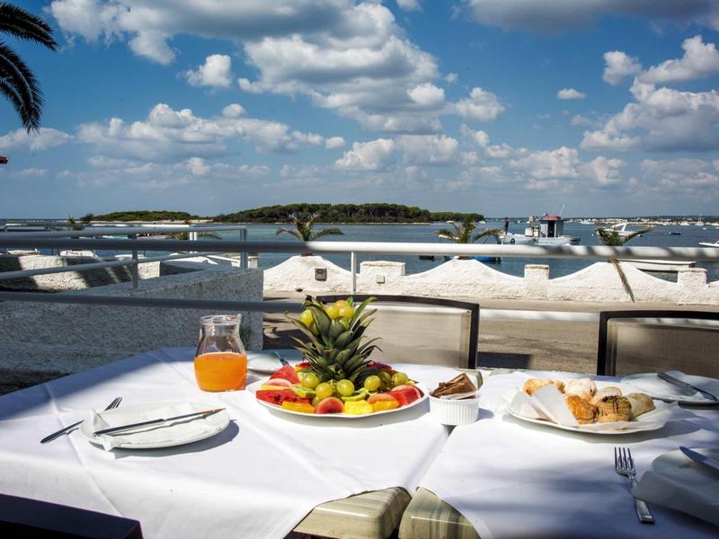 Tisch mit Obst, Saft und Gebäck am Wasser, viele Boote im Hafen unter blauem Himmel.