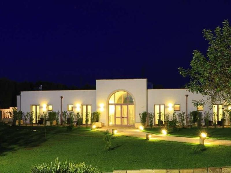 Ein beleuchtetes, weißes Gebäude mit Bogenfenstern, umgeben von Bäumen und grünem Rasen bei Nacht.