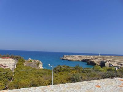 Küste mit blauem Meer, grünem Buschwerk und bewölktem Himmel, felsige Landzunge und Straßenlaternen.