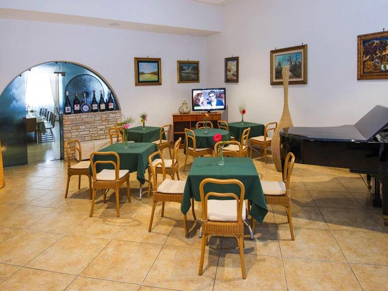 Großer Raum mit Tischen, Stühlen, grünem Stoff, Klavier und Wandbildern, heller Bodenfliese.