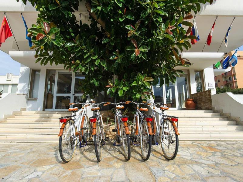 Fünf Fahrräder lehnen vor einer Treppe unter einem großen grünen Baum, Gebäude im Hintergrund.