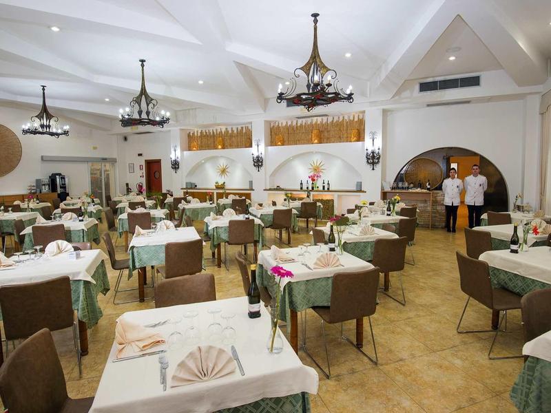 Großer heller Speisesaal mit weißen Tischtüchern, grünen Läufern und zwei stehenden Kellnern.