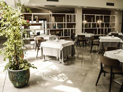 Helles Restaurant mit weißen Tischtüchern, schwarzen Stühlen und großer Pflanze im vorderen Bereich.