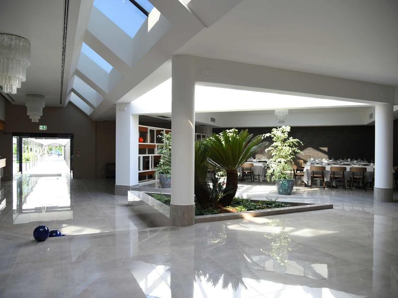 Großer heller Raum mit Glasdach, Pflanzeninsel in der Mitte und Tisch mit Stühlen im Hintergrund.