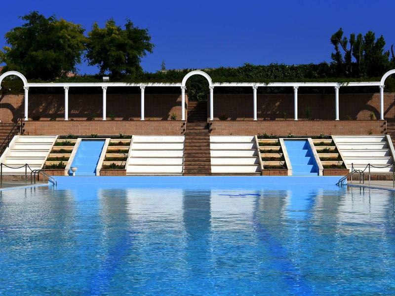 Leerer Schwimmbeckenbereich mit blauer Wasserfläche, weißen Liegestühlen und Bäumen im Hintergrund.