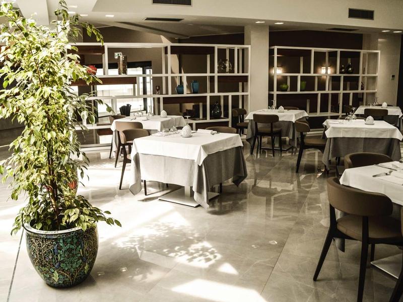 Helles Restaurant mit weißen Tischtüchern, schwarzen Stühlen und großer Pflanze im vorderen Bereich.