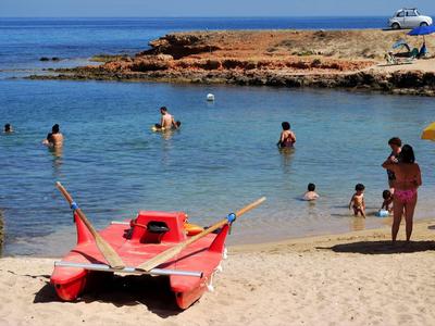 Strand mit rotem Tretboot im Vordergrund, Menschen schwimmen und spielen im klaren blauen Wasser.