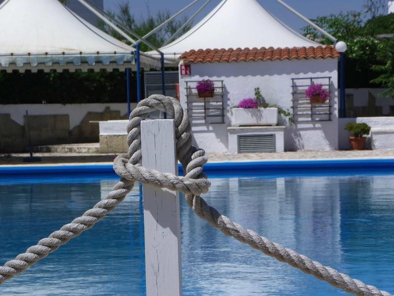 Blauer Pool mit Seil, das an weißem Pfosten befestigt ist, im Hintergrund Gartenhaus und Sonnensegel.