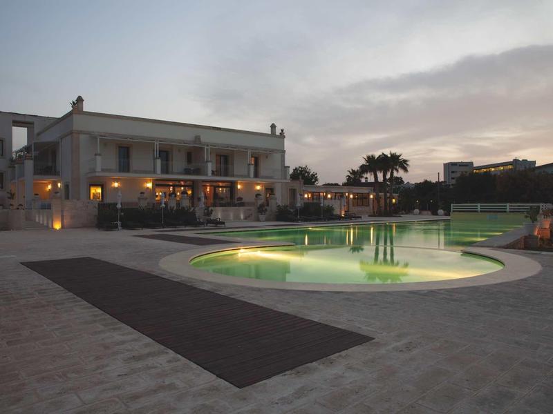 Moderne Villa mit beleuchtetem Pool, großzügiger Terrasse und Palmen im Abendlicht.