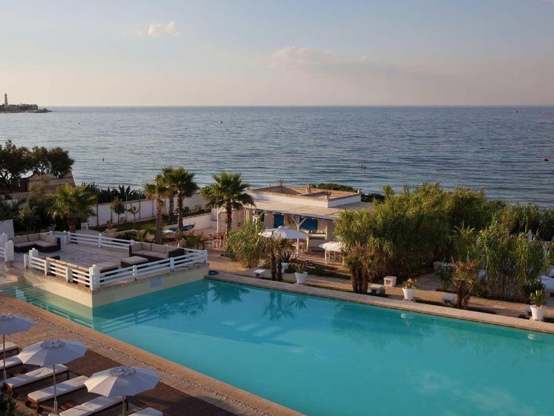 Ein Pool mit Liegen am Strand, Palmen und Meer im Hintergrund bei klarem Himmel.
