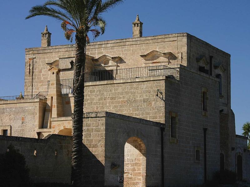 Gebäude aus hellem Stein mit Bogen und Palmen vor blauem Himmel an sonnigem Tag.