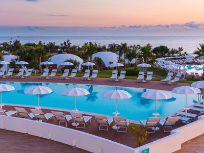 Hotelpool mit Liegestühlen, Sonnenschirmen, Palmen und Meer im Hintergrund bei Sonnenuntergang.