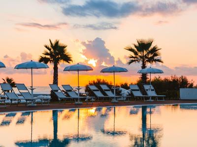 Schwimmbad mit Liegestühlen und Sonnenschirmen, Palmen und buntem Sonnenuntergang im Hintergrund.