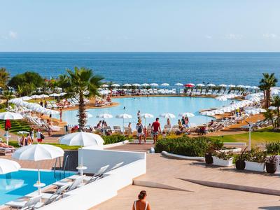 Großer Pool mit Sonnenschirmen und Liegestühlen, Palmen und Meer im Hintergrund unter blauem Himmel.