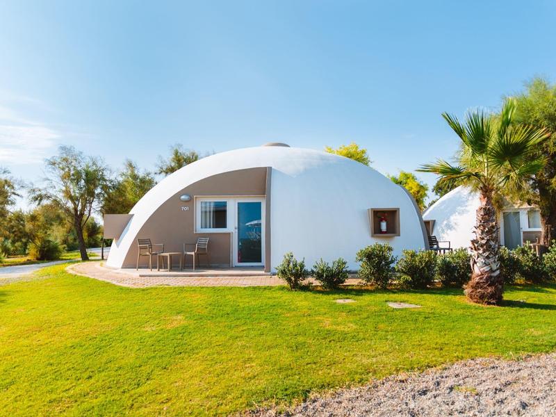 Weißes, kuppelförmiges Haus mit Terrasse, umgeben von grünem Rasen und Palmen.
