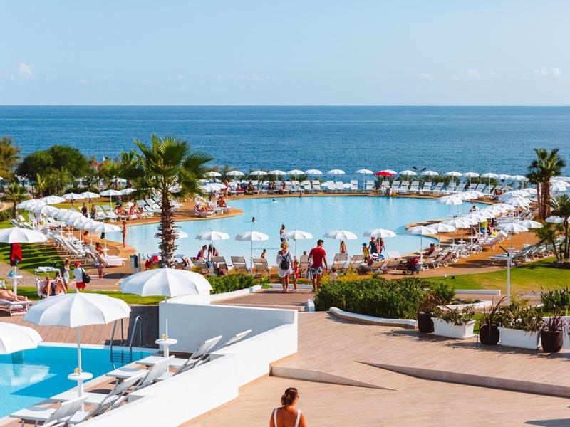 Großer Pool mit Sonnenschirmen und Liegestühlen, Palmen und Meer im Hintergrund unter blauem Himmel.