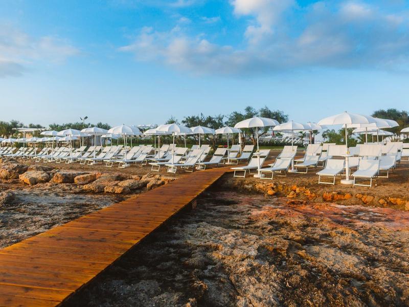 Ein Holzsteg führt zu Reihen von weißen Liegestühlen und Sonnenschirmen am steinigen Strand.