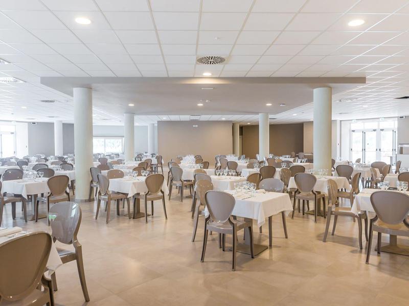 Großer heller Speisesaal mit vielen rechteckigen Tischen und grauen Stühlen, hoher Decke.