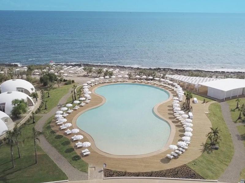 Großer, geschwungener Pool umgeben von weißen Liegestühlen und Kuppelbungalows am Meer.