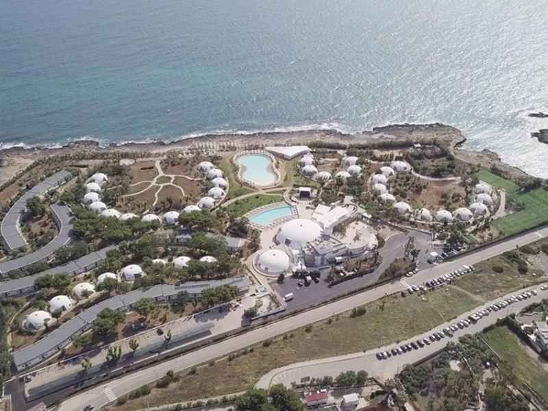 Luftaufnahme einer Küstenanlage mit weißen Gebäuden, Pools und Umzäunung nahe blauem Meer.