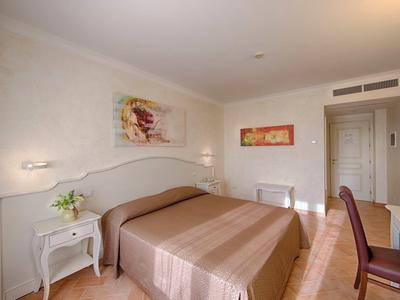 Helles Schlafzimmer mit großem Bett, Nachttischen, Stuhl, Wandbildern und Deckenbeleuchtung.