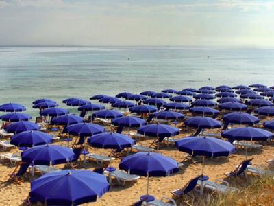 Viele blaue Sonnenschirme und Liegestühle an einem ruhigen Sandstrand am Meer.