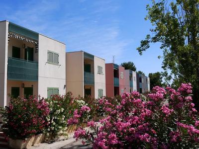 Mehrfamilienhäuser mit Balkonen in Pastellfarben, davor blühende rosa Sträucher und blauer Himmel.