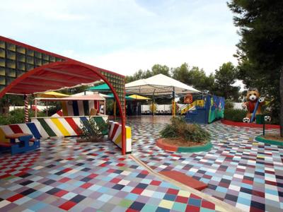 Bunter Spielplatz mit Mosaikboden, roten Sitzbänken, Zelt und grünen Bäumen im Hintergrund.