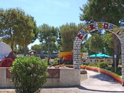 Park mit Bäumen, bunter Steinbogen und Sitzgelegenheiten an einem sonnigen Tag.