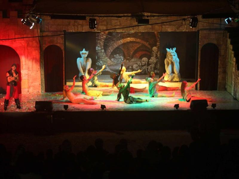 Bühnenbild mit farbigen Figuren und Schauspielern vor einer Höhlenkulisse unter warmer Beleuchtung.