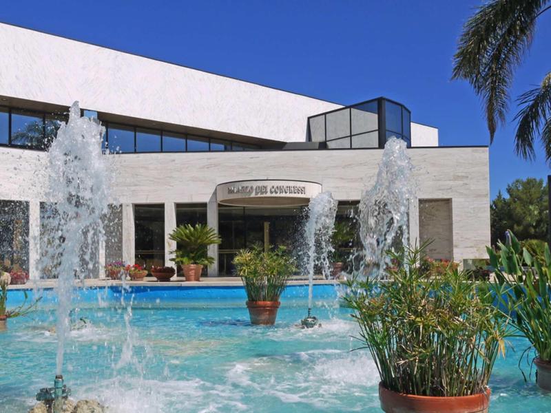Modernes Gebäude mit Springbrunnen und klarem Wasser vor blauem Himmel und grünen Pflanzen.