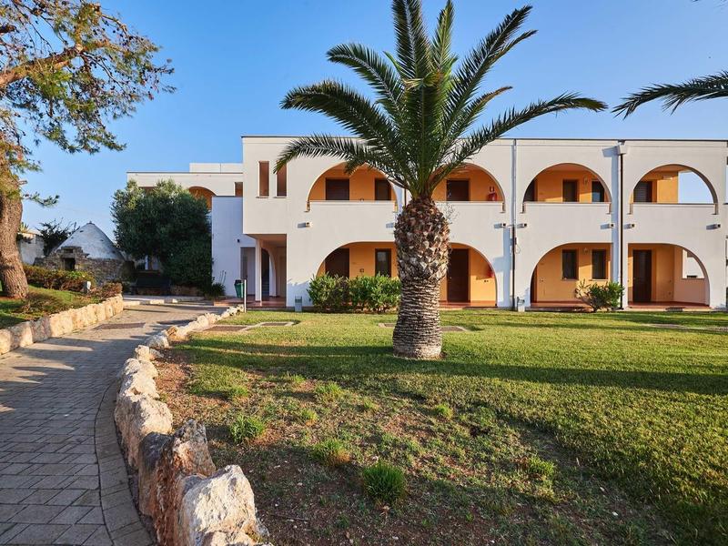 Zweistöckiges weißes Haus mit Bögen, Palmen und grünem Rasen unter blauem Himmel.