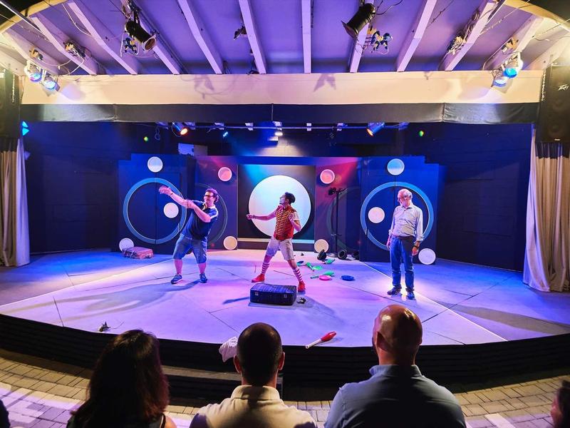 Bühne mit drei Tänzern, buntem Hintergrund und Publikum im Vordergrund. Bühne in warmem Licht.