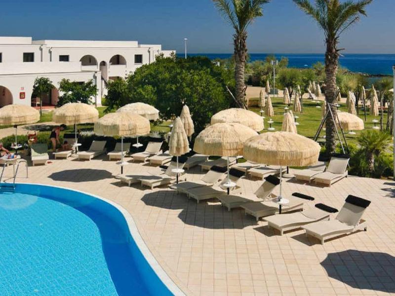 Großer blauer Pool mit Liegestühlen und Sonnenschirmen, Palmen und Blick aufs Meer.