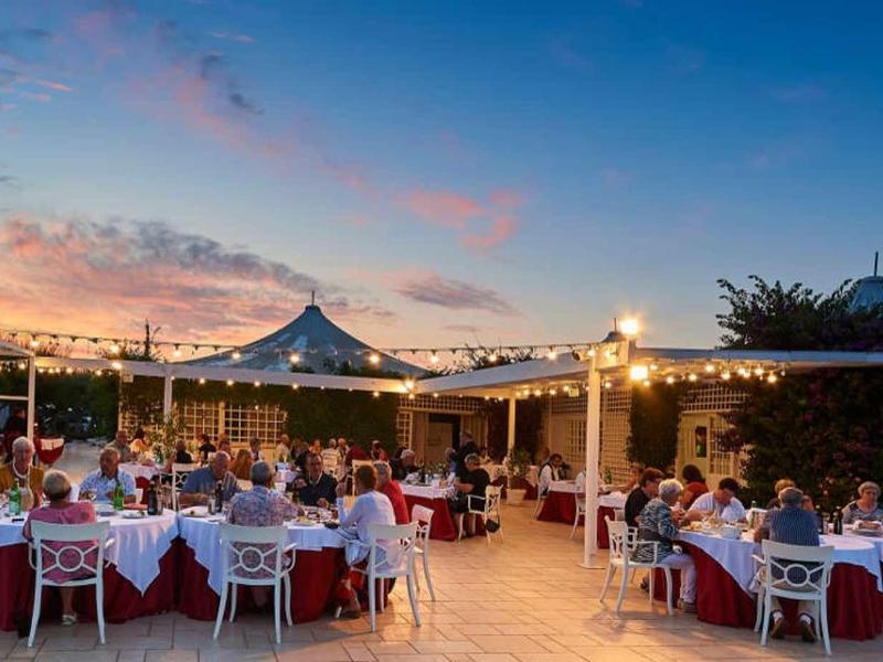 Draußen festlich gedeckte Tische mit weißen Hussen, Lichterketten und Menschen bei Sonnenuntergang.