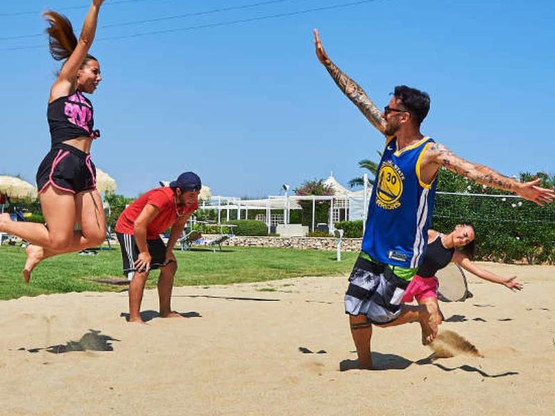 Vier Personen spielen Beachvolleyball, zwei springen hoch, Sand und blauer Himmel im Hintergrund.
