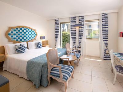 Helles Hotelzimmer mit Doppelbett, blauen Akzenten und Sitzbereich vor großen Fenstern.