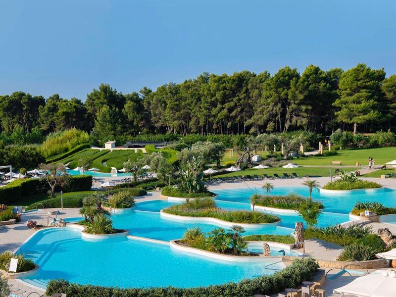 Großer Hotelpool mit mehreren Becken und Sonnenliegen, im Hintergrund Bäume und blauer Himmel.