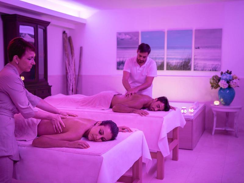 Zwei Frauen erhalten eine Massage in einem lila beleuchteten Spa-Raum mit Meerblick-Kunst an der Wand.