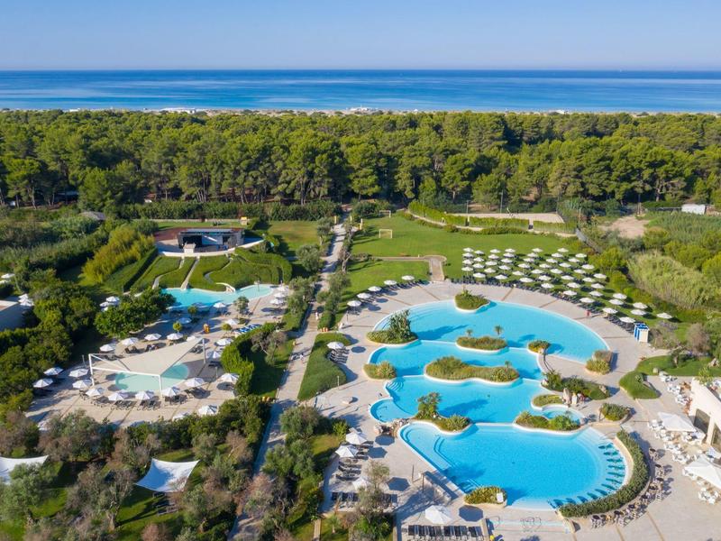 Großer Hotelpool mit Inseln, Sonnenliegen und grüner Umgebung nahe dem Meer.