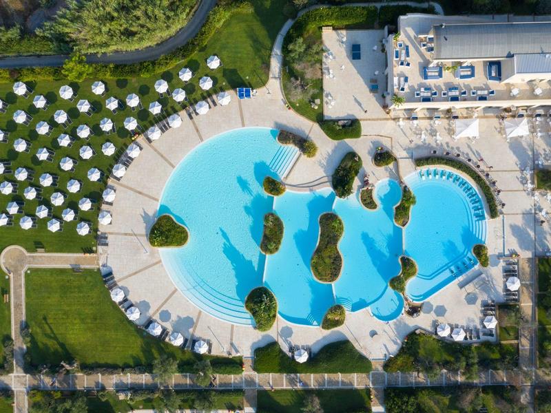 Luftaufnahme eines großen Hotelpools mit Sonnenschirmen und Liegestühlen auf einer grünen Wiese.