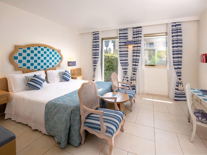 Helles Hotelzimmer mit Doppelbett, blauen Akzenten und Sitzbereich vor großen Fenstern.